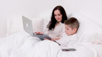 mom and son under blanket are sitting on soft bed in the hands of boy tablet playing online freelancer on laptop spend time with son during work recover stay at home watch movie online communication video