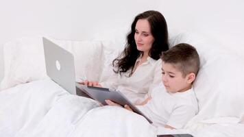 mom and son under blanket are sitting on soft bed in the hands of boy tablet playing online freelancer on laptop spend time with son during work recover stay at home watch movie online communication video