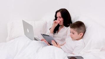 woman coughing mom and son under blanket are sitting on soft bed boy tablet playing online freelancer on laptop spend time with son during work recover stay at home watch movie online communication video