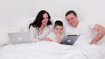 husband wife in bed with little son looking at tablet cartoon interesting game pictures laughing talking communication real people at home love life joyful people space for text ad social relations video