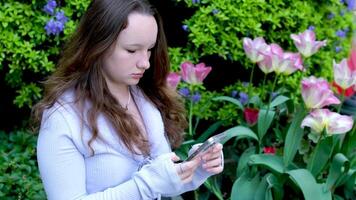 Jeune fille en marchant dans parc et parlant par téléphone intelligent près arbre dans fleurir. concept de moderne La technologie et printemps dans la nature. video