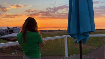 a teenage girl in Green T-shirt stands with her back to the camera on balcony and shoots the sunset vacation in a hotel on the sea or ocean beautiful view of nature landscape space for text background video