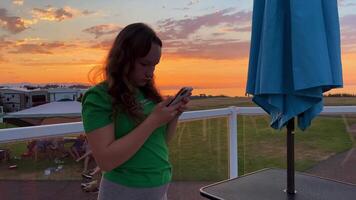 un Adolescente niña con rubio rojo pelo en un verde camiseta es en pie en el hotel balcón por el mar naranja horizonte soleado puesta de sol noche ella es en pie y mirando a el teléfono en social redes video