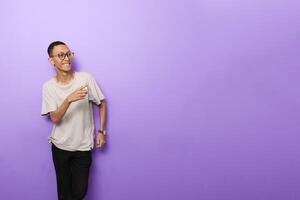 an Asian man in a plain white t-shirt with glasses gesturing to something to the side isolated on purple background photo