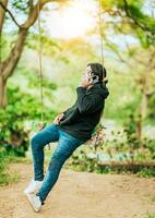 joven hombre sentado en un columpio vocación en el teléfono en un jardín. contento hombre balanceo hablando en el teléfono foto