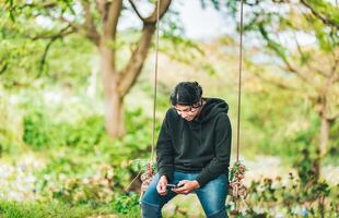 Smiling guy sitting on a swing with smart phone. Young man sitting on a swing using cell phone in nature photo