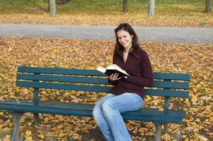 joven mujer leyendo libro sentado en parque banco en otoño foto