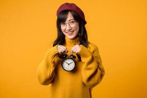 A young Asian woman, wearing a yellow sweater and red beret, smiles as she holds an alarm clock against a vibrant yellow background. photo