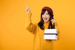 Stylish young Asian woman in her 30s, holding a shopping paper bag and pointing her finger, dressed in a yellow sweater and red beret against a yellow background. photo