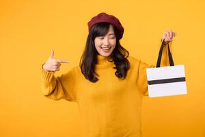 Young Asian woman in her 30s, holding a shopping paper bag, pointing her finger with a cheerful expression, wearing a yellow sweater and red beret against a yellow background. photo