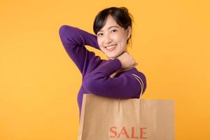 un Encantado joven mujer, vistiendo un púrpura camisa y participación un compras bolso en contra un amarillo fondo, simboliza el emoción de compras y hallazgo fantástico descuentos foto