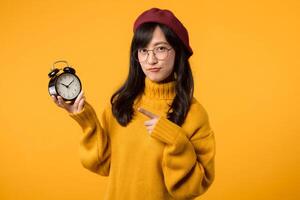 A punctual young Asian woman, wearing a yellow sweater and red beret, points to her alarm clock, ensuring she's always on time, against a vibrant yellow background. photo