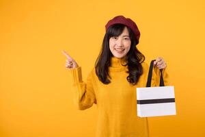 joven asiático mujer en su 30, con un elegante instinto, participación un compras papel bolsa, señalando su dedo, poniéndose un amarillo suéter y rojo boina en contra un amarillo antecedentes. foto