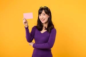 Wearing purple shirt and sunglasses against a vibrant yellow backdrop, a lady displays a blank card board, ready for your message or advertisement. photo