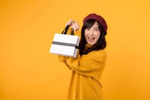 Young Asian woman in her 30s, with a shopping paper bag, displaying elegance in a yellow sweater and red beret during her shopping spree against a yellow background. photo