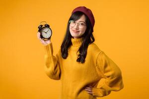 In a stylish scene, a young woman in a yellow sweater and red beret confidently holds her hip next to an alarm clock, showcasing punctuality and fashion sense. photo