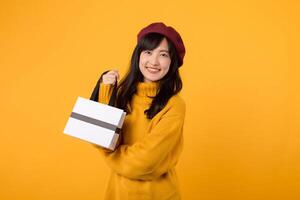 joven asiático mujer en su 30, que lleva un compras papel bolsa, vistiendo un amarillo suéter y rojo boina, disfrutando un exitoso compras día en contra un amarillo antecedentes. foto