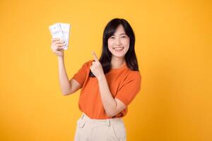 Dynamic young Asian woman in her 30s, pointing finger to cash money dollars, standing on vibrant yellow backdrop. photo
