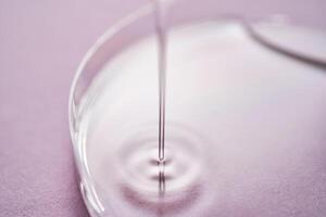 Serum or cosmetic oil flows into a transparent bowl on a purple background. photo