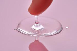 A finger touches a drop of cosmetic product on a pink background. photo