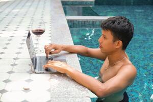 asiático hombre con un vaso de rojo vino mirando contento mientras comprobación correo, noticias, social redes, trabajando en computadora portátil, escritura Blog, estudiando a hogar, complejo, hotel en fin de semana. en línea Lanza libre concepto. foto