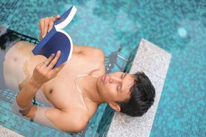 asiático hombre tendido en el nadando piscina leyendo un libro en el verano en un soleado día. relajante en un patio interior nadando piscina para relajarse en verano vacaciones día. foto