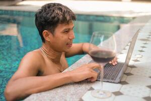 asiático hombre con un vaso de rojo vino mirando contento mientras comprobación correo, noticias, social redes, trabajando en computadora portátil, escritura Blog, estudiando a hogar, complejo, hotel en fin de semana. en línea Lanza libre concepto. foto