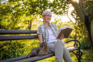 contento mayor mujer disfruta leyendo libro y gasto hora con su gato mientras sentado en un banco en su jardín. foto