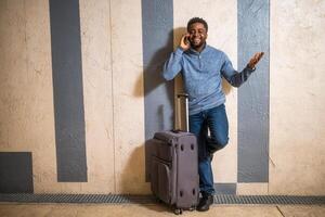 Happy man with suitcase using phone while leaning against a wall in passage of the railway station. Copy space on image for your advert or text. photo