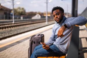 Angry man with a suitcase and mobile phone sitting on a bench at the railway station photo