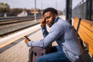 Angry man with a suitcase and mobile phone sitting on a bench at the railway station. photo