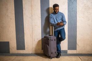 Happy man with suitcase using phone while leaning against a wall in passage of the railway station. Copy space on image for your advert or text. photo