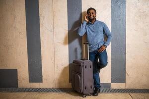 Happy man with suitcase using phone while leaning against a wall in passage of the railway station. Copy space on image for your advert or text. photo