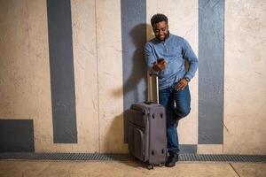 Happy man with suitcase using phone while leaning against a wall in passage of the railway station. Copy space on image for your advert or text. photo