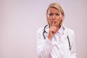 Portrait of mature female doctor showing silence sign on gray background. photo