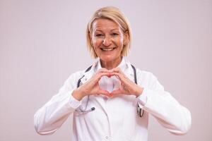 Portrait of mature female doctor showing heart shape with hands on gray background. photo