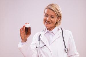 Portrait of mature female doctor holding bottle of pills on gray background. photo