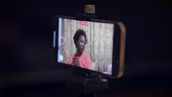 A woman is filming a makeup tutorial on her smartphone, likely using a tripod for vlogging video