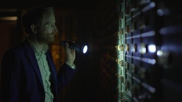 Man in a suit uses a flashlight to explore a vault in a dimly lit room filled with storage boxes video