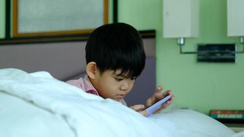 en liten asiatisk pojke lögner på de säng spelar med hans telefon. video