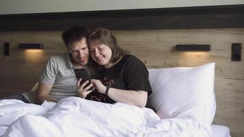 jovem casado casal com sorrisos olhando às Smartphone tela juntos enquanto sentado em cama. jovem mais Tamanho mulher e delgado homem dentro quarto escolher Comida Entrega para seus lar. video