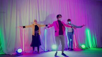 Three people dancing at a party with colorful lights and a white backdrop, wearing casual clothes and face masks video