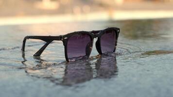 Sunglasses left on shore. A black sunglasses near a pool with waves reflection on pebbles under sun rays. Active Resort Relax, Carefree Living, Freedom Enjoyment Outdoors video