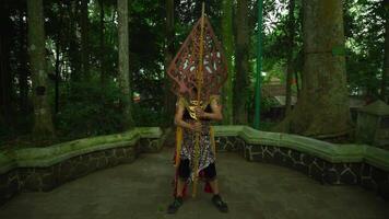 Two performers in traditional Javanese costumes and masks, posing in a serene forest setting with a cultural sculpture video
