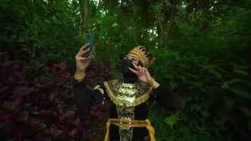 la personne dans traditionnel ethnique tenue et masque en utilisant une intelligent téléphone dans une luxuriant vert forêt réglage video