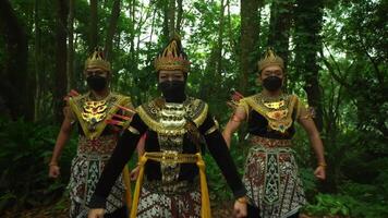 Three performers in traditional Balinese costumes and masks stand in a lush forest, their expressions obscured by black face masks video