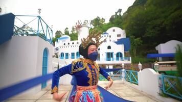 A person in vibrant traditional clothing and a mask performs a dance with blue ribbons, set against a whimsical backdrop video