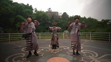 Three performers in clown makeup and whimsical costumes dancing joyfully outdoors with a lush, green park and a building video