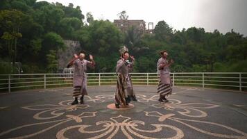 Three performers in traditional costumes and masks dancing outdoors with a lush green backdrop and a historic building video