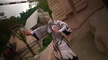 Performer in traditional costume and mask dancing in front of a large stone sphinx statue, with lush greenery and wooden buildings video